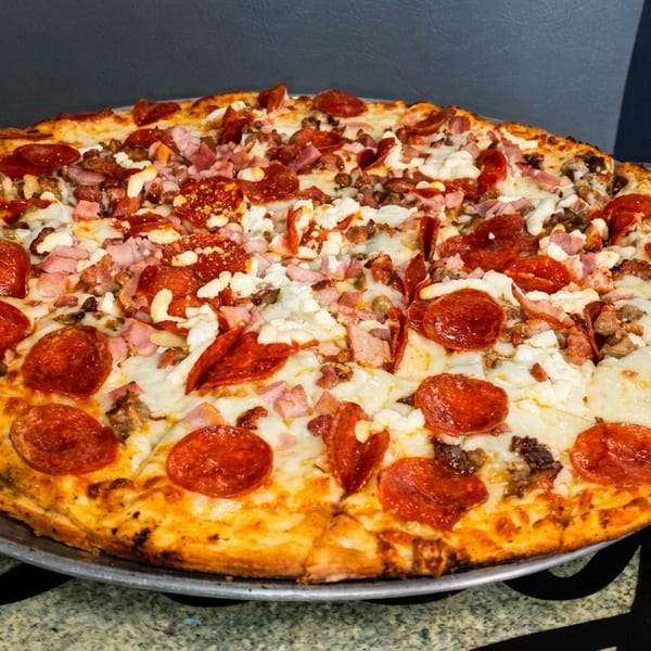 Large, freshly baked pizza with pepperoni, ham, and cheese on a metal pan.
