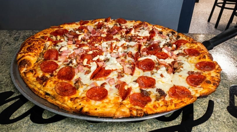 Large, freshly baked pizza with pepperoni, ham, and cheese on a metal pan.