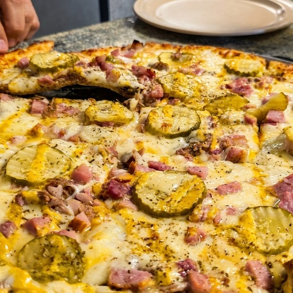 Hands serving a slice of pizza topped with pickles, ham, and melted cheese from a large pie.