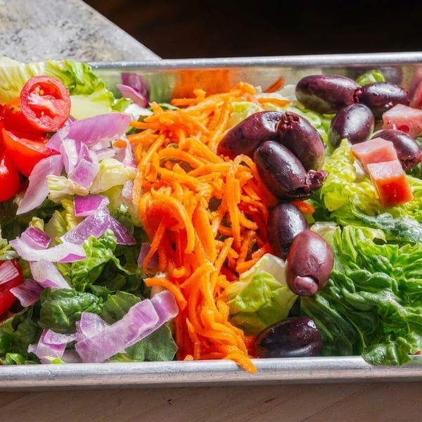 Fresh salad with lettuce, tomatoes, red onions, shredded carrots, olives, and diced ham in a metal tray.