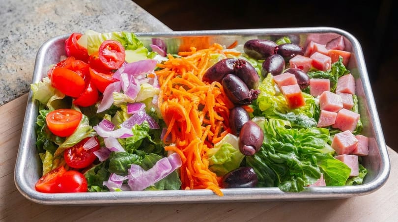 Fresh salad with lettuce, tomatoes, red onions, shredded carrots, olives, and diced ham in a metal tray.