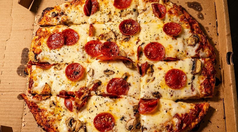 Square-cut pepperoni and mushroom pizza with melted cheese and herbs in a cardboard box.