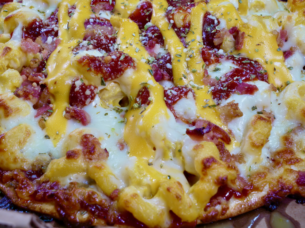 A close up of Pork Mac Attack pizza that has a garlic butter base, pork, mac & cheese and bacon.