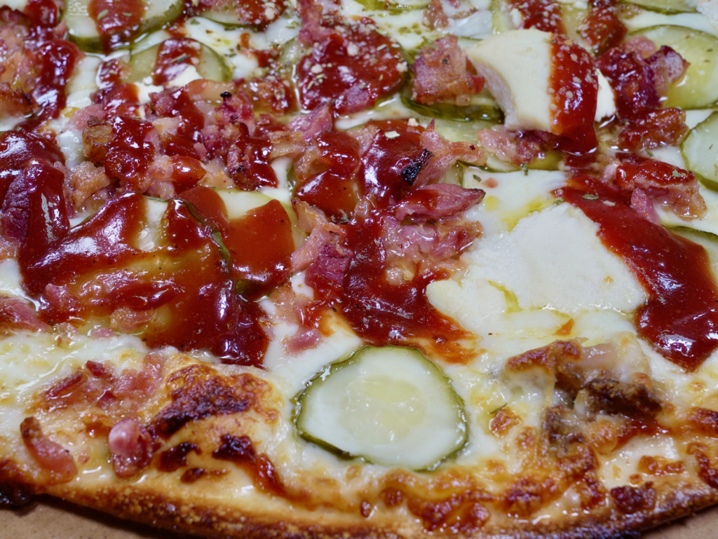 A close up of The Mechanic pizza that has a garlic butter BBQ base, pork, bacon, pickles, and ricotta cheese.