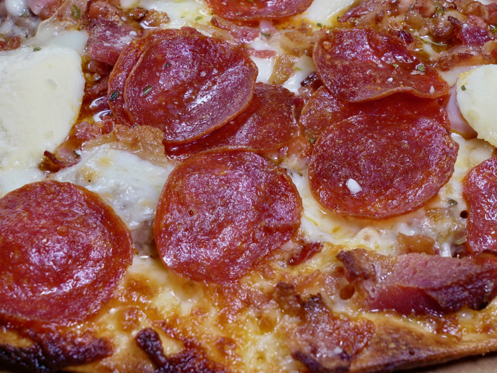 A close up of The Walter White pizza that has a garlic butter base, sausage, pepperoni, ham, bacon, and ricotta cheese.